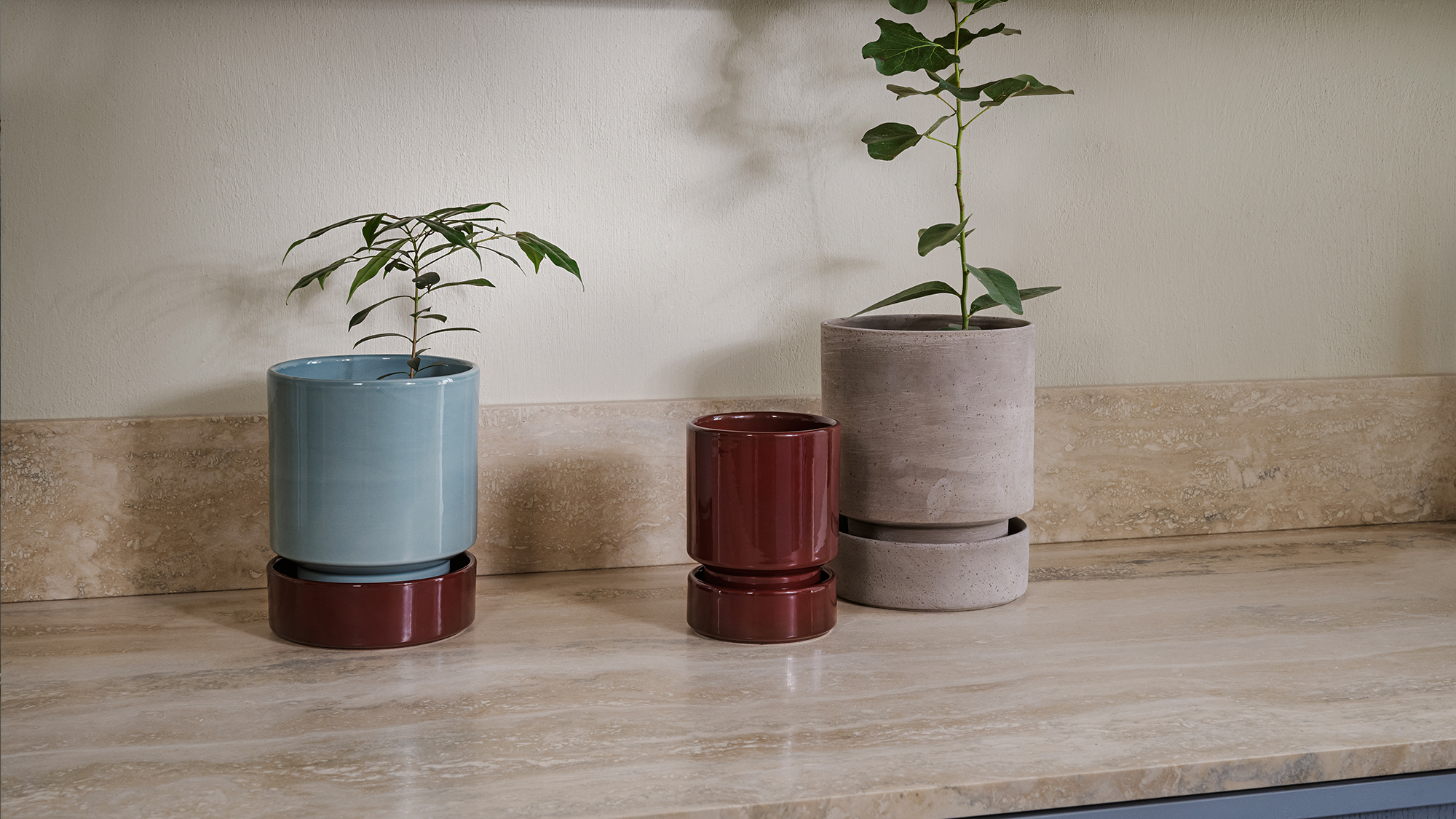 Bergs Potter Hoff pots in misty blue with deep burgundy saucer, deep burgundy, and raw grey terracotta with matching saucer, styled on a marble countertop.