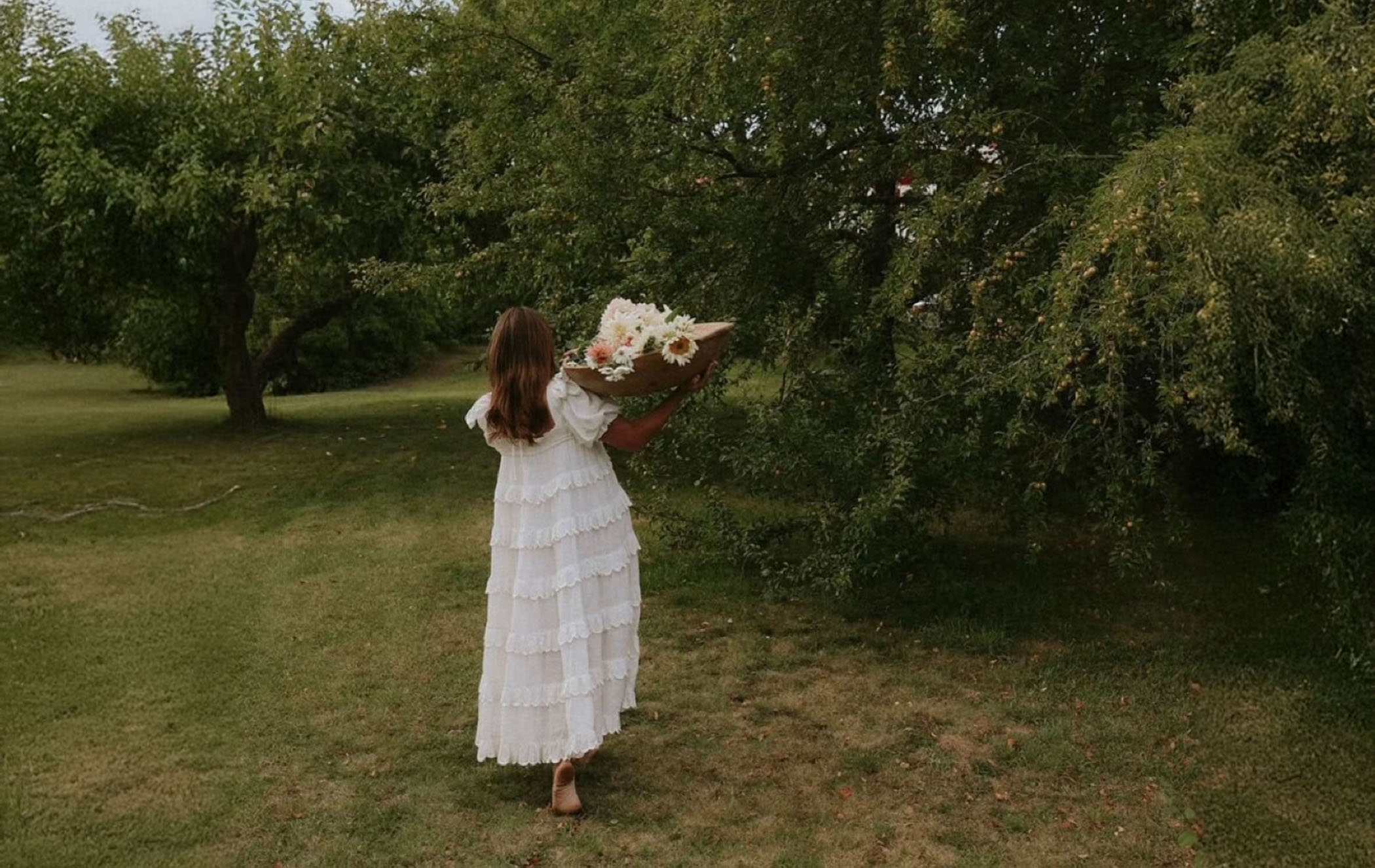 Woman in white dress carrying flowers through orchard