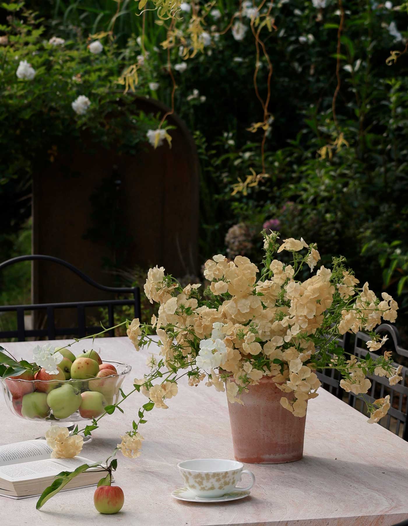 Emilia pot in Rosa terracotta Ø21 styled with pale garden flowers on an outdoor table with apples and a teacup.