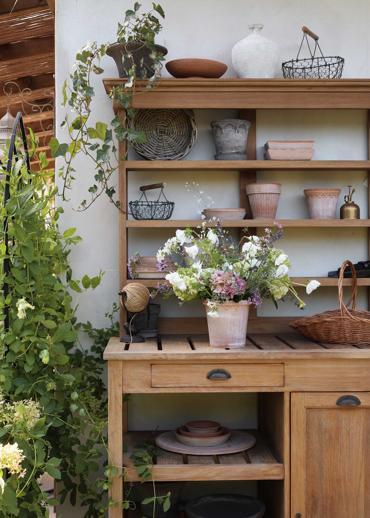 Emilia pot in Rosa terracotta filled with a mixed garden bouquet, styled on a wooden potting bench surrounded by terracotta pots, baskets and climbing plants.