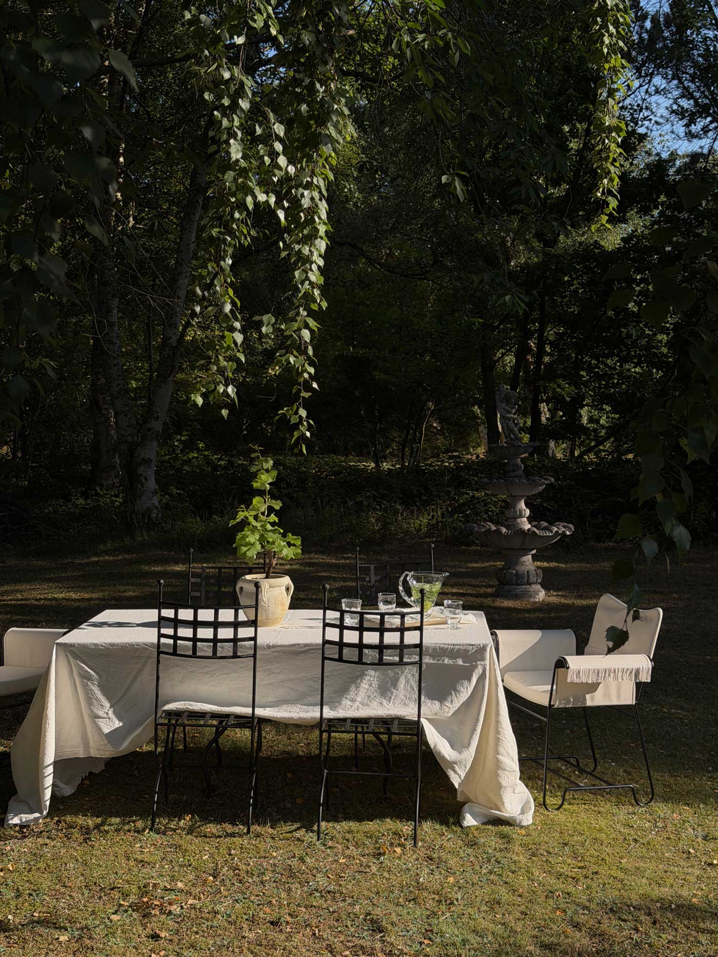 Woman in a white dress setting an outdoor dining table in the garden of a Danish summer house, surrounded by trees and potted plants.