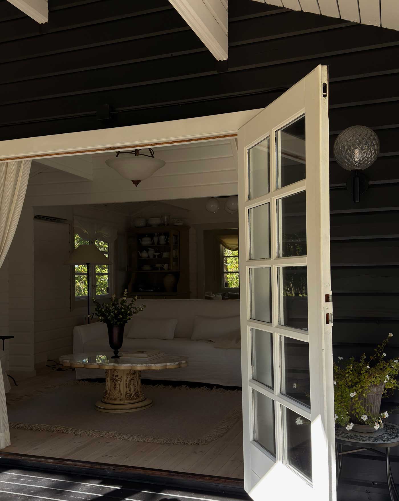 Open white double doors leading from the terrace into a light-filled Danish summer house living room with natural materials and soft neutral tones.