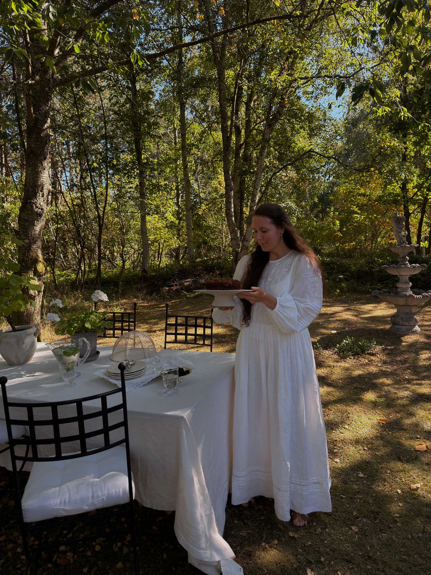 Woman in a white dress setting an outdoor dining table in the garden of a Danish summer house, surrounded by trees and potted plants.