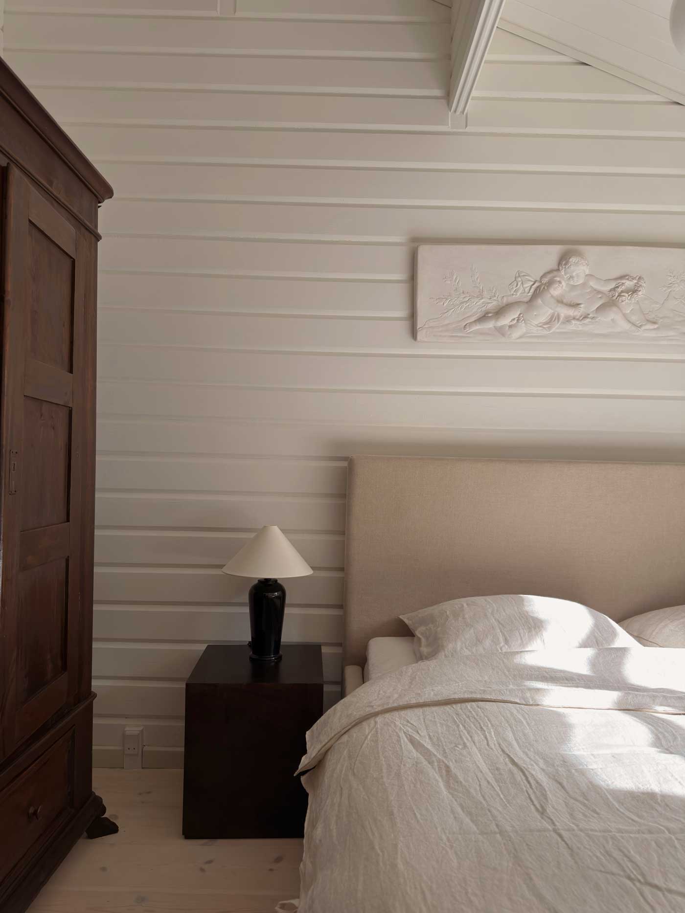 Light-filled bedroom in a Danish summer house with white wood-panelled walls, natural linen bedding and dark wooden furniture.