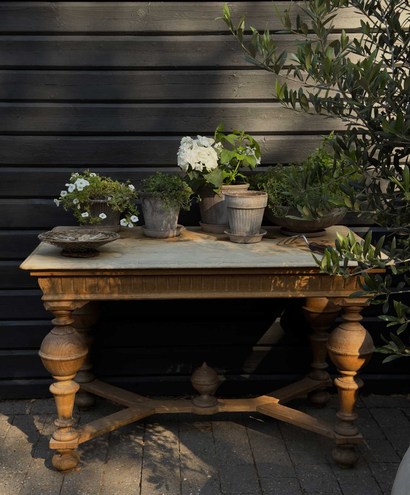 Outdoor dining area beside a dark wooden Danish summer house, styled with white-framed windows, terracotta pots and green plants.