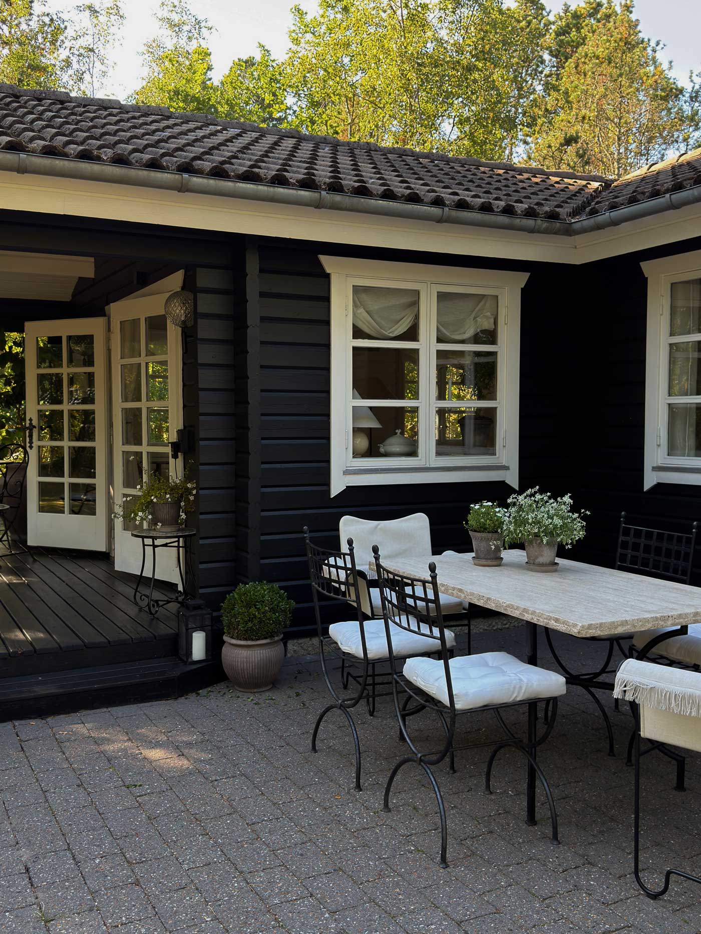 Outdoor dining area beside a dark wooden Danish summer house, styled with white-framed windows, terracotta pots and green plants.